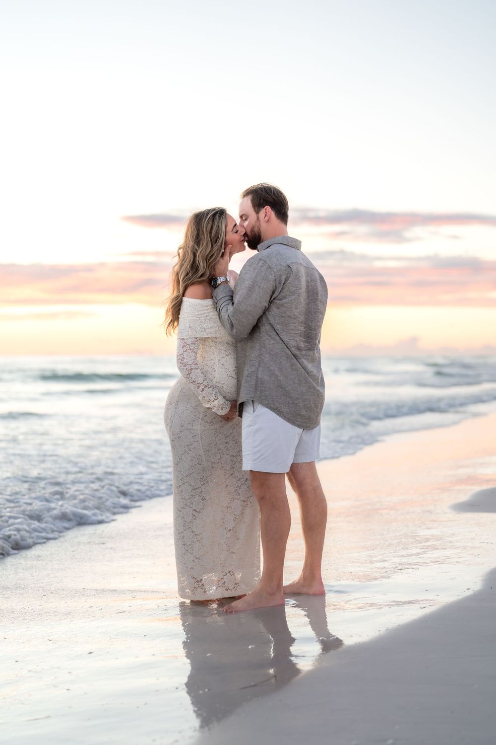 Expecting couple sharing tender moment barefoot on beach at sunset, woman in white lace maternity gown.