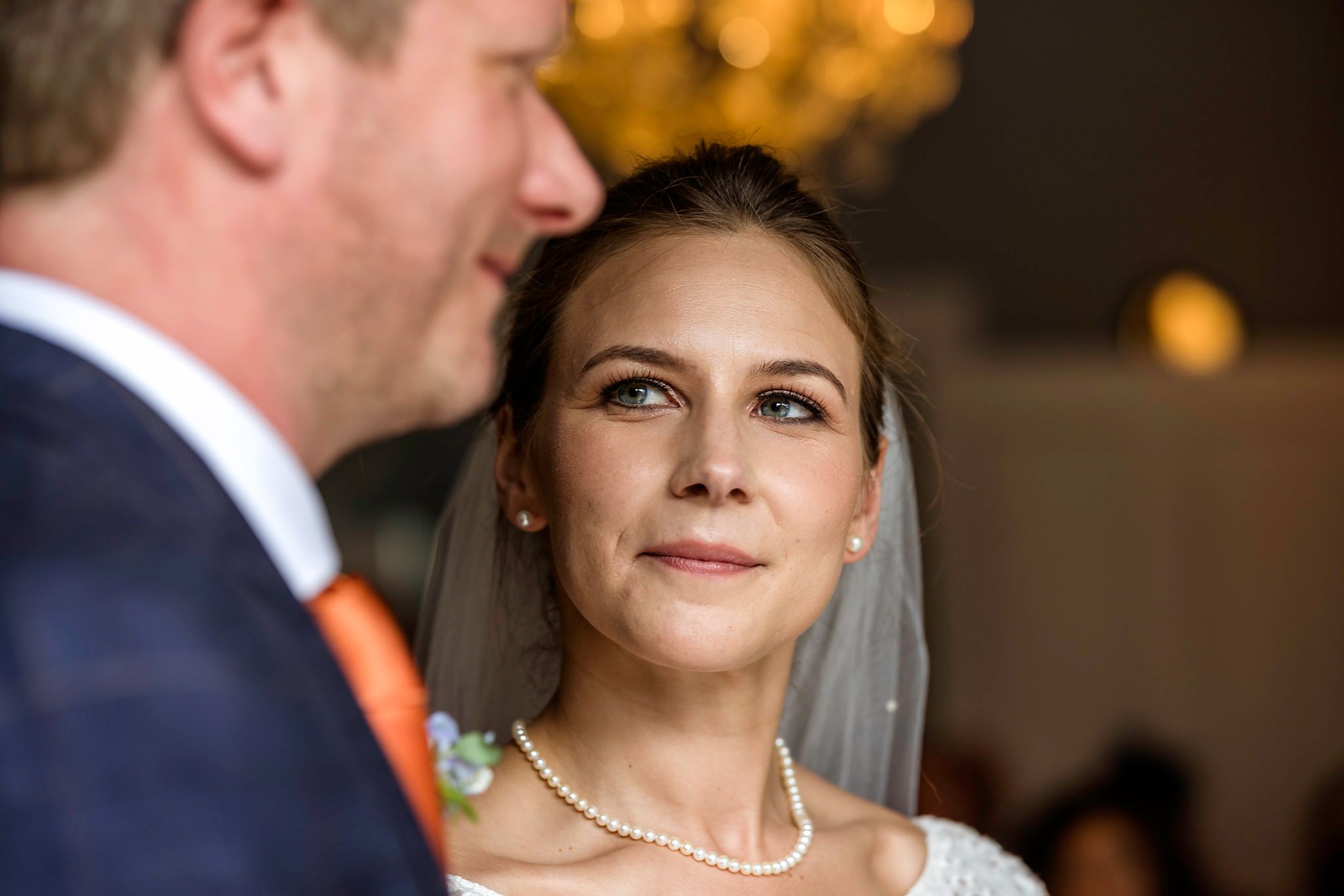 Bride in pearl necklace and veil gazes lovingly at groom in orange tie during wedding ceremony.