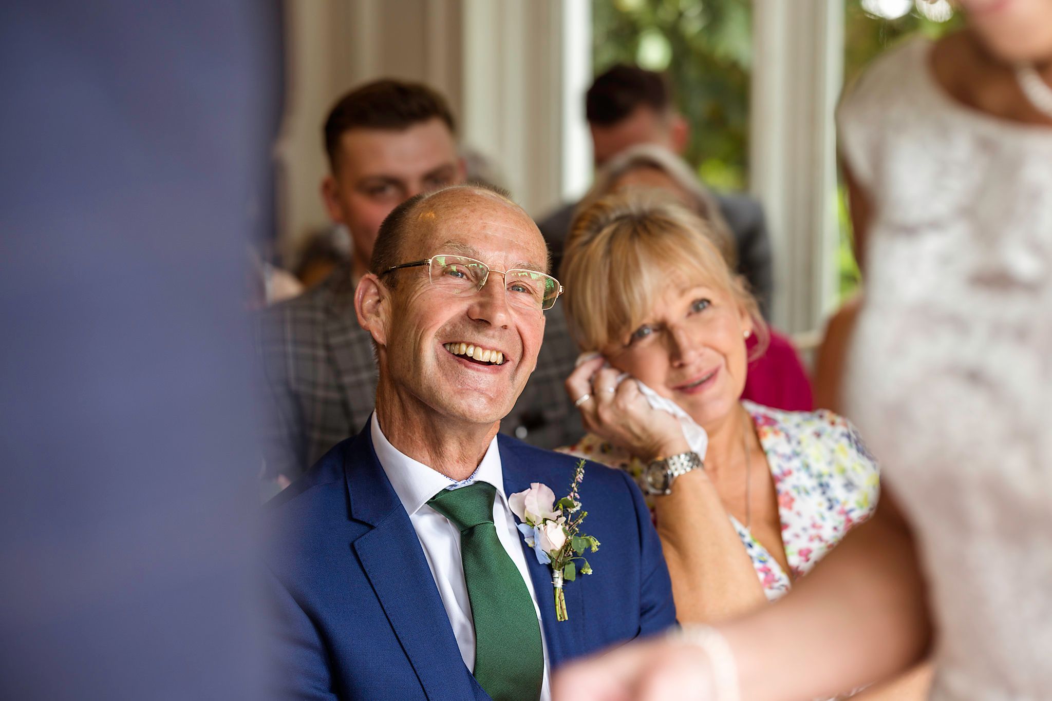 Joyful older couple at a wedding, man in blue suit with green tie laughing, woman smiling emotionally in floral dress.