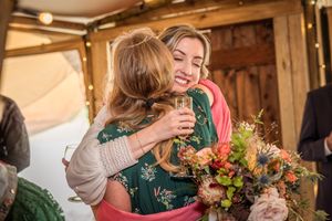 Two women sharing a warm hug at a wedding reception, one holding champagne and a colorful bouquet.