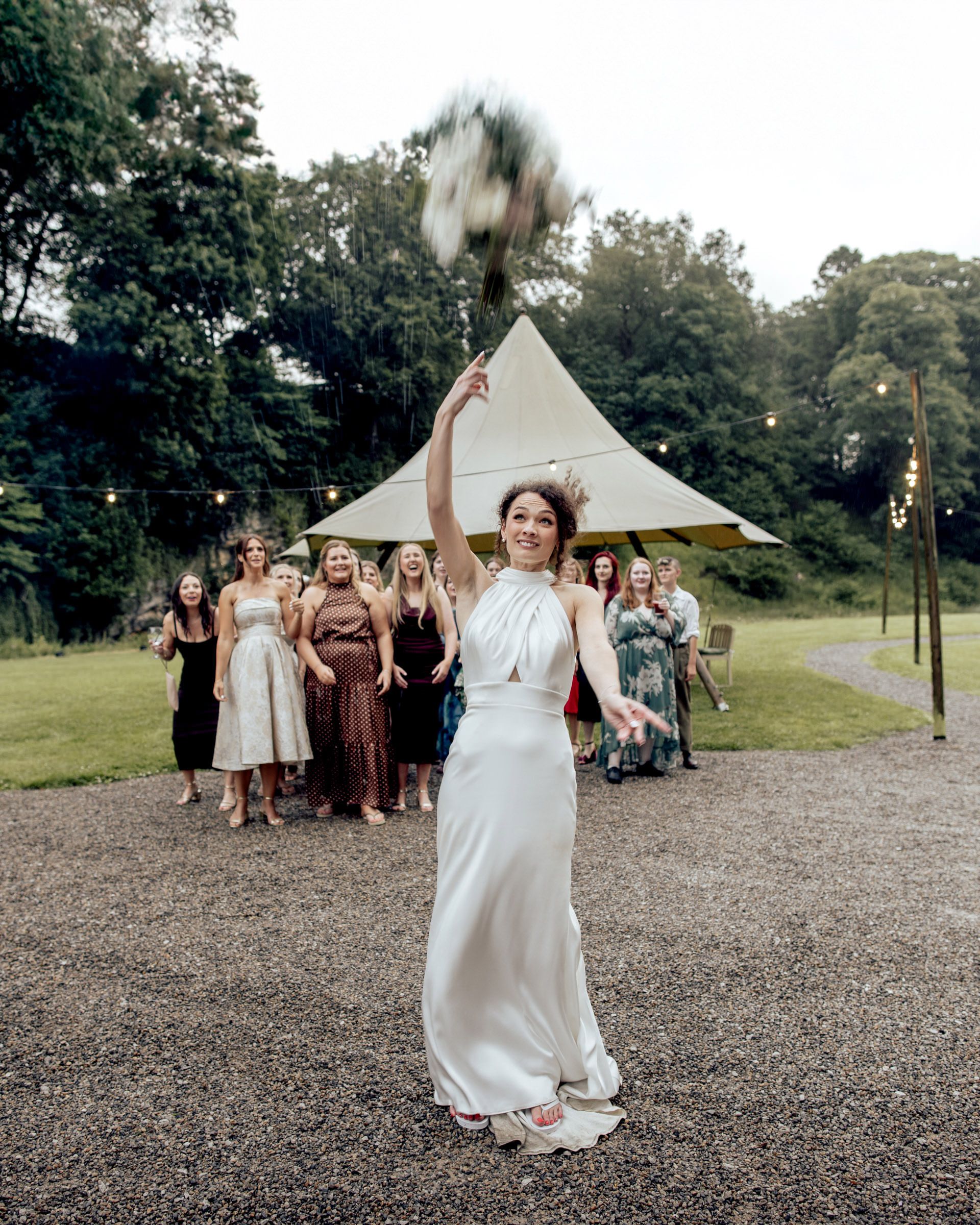 Bride in white gown tossing bouquet to bridesmaids outdoors near a tent with string lights at a wedding reception.