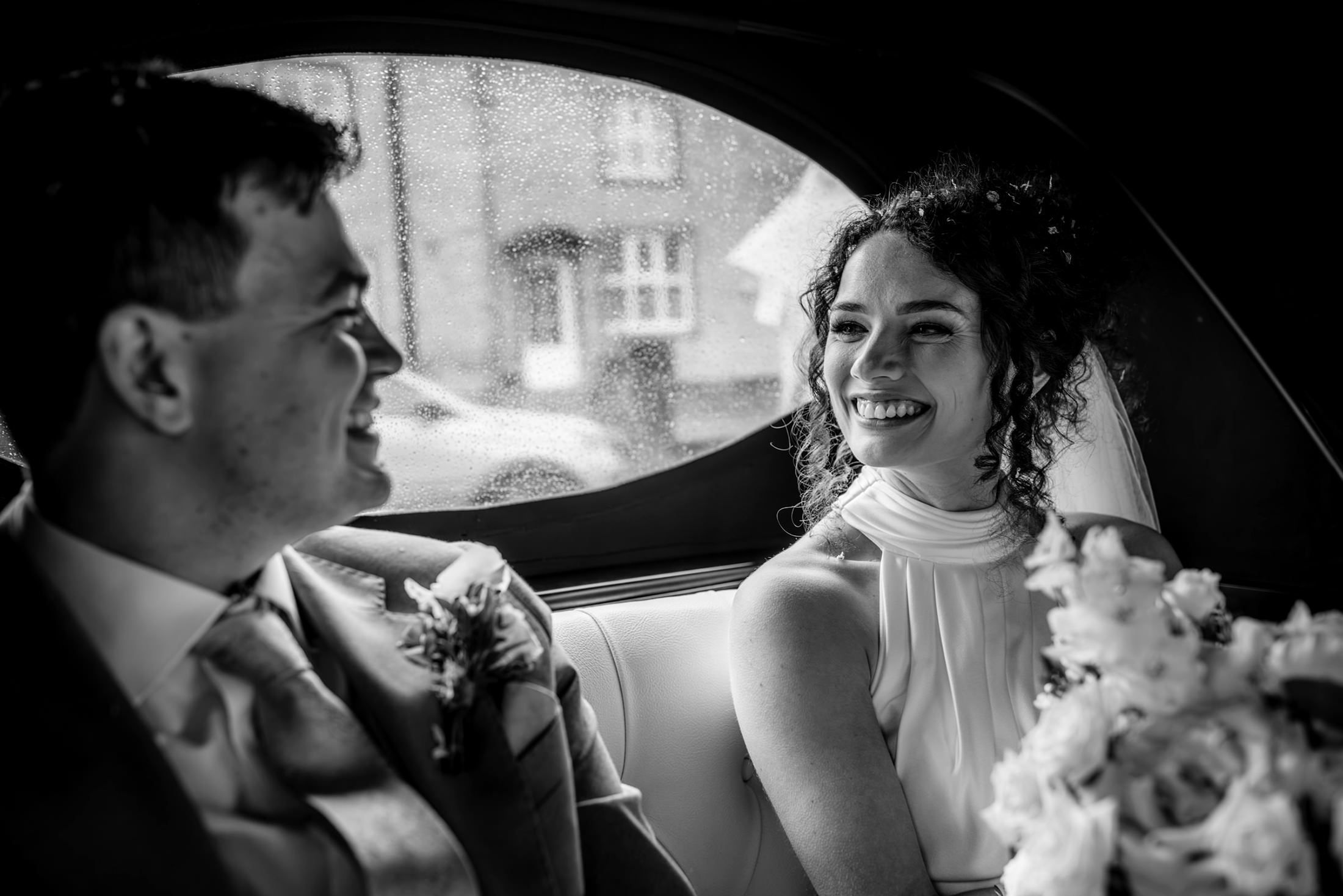 Black and white photo of a smiling bride and groom sitting together in a car on their wedding day.