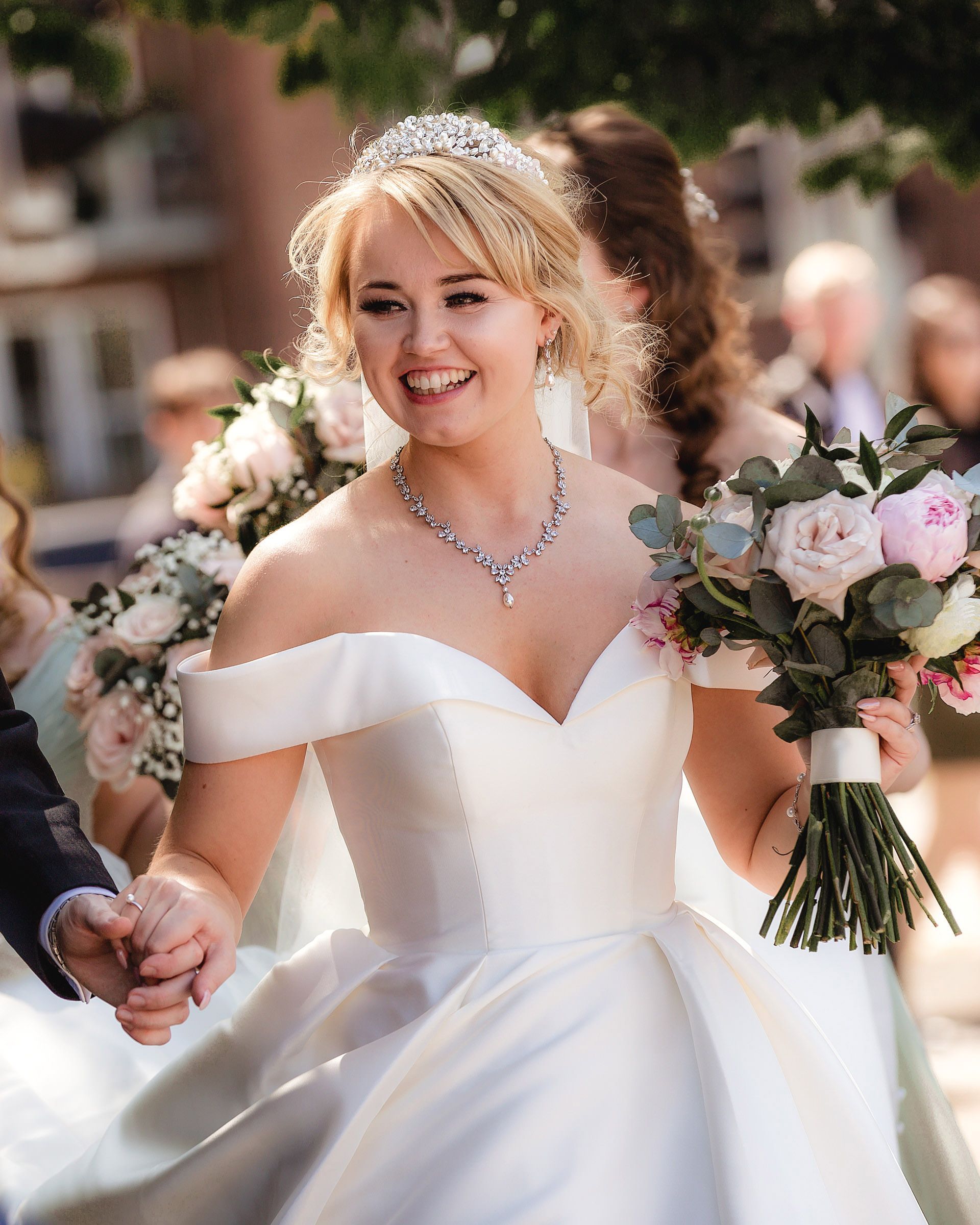 Smiling bride in off-shoulder white gown, tiara, and diamond necklace holds pink bouquet outdoors.