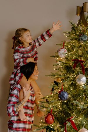 Family in matching plaid pajamas decorating a Christmas tree with ornaments and lights.