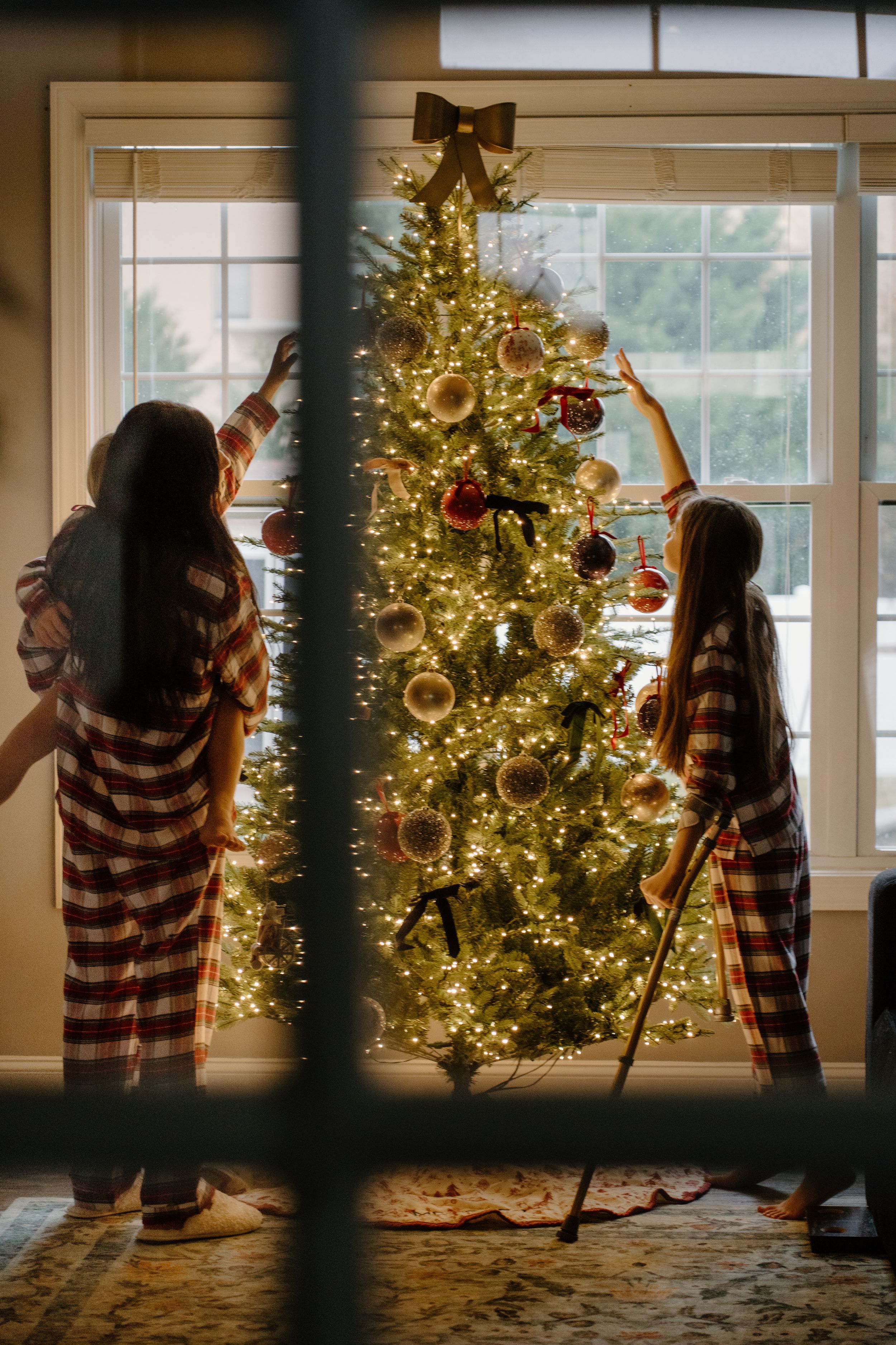 Two people in matching striped pajamas decorate a glowing Christmas tree with ornaments near a bright window.