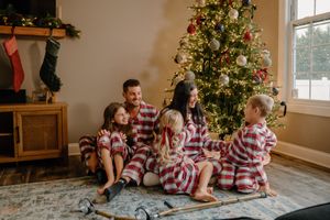 Family in matching striped pajamas sits together by decorated Christmas tree opening presents on Christmas morning.