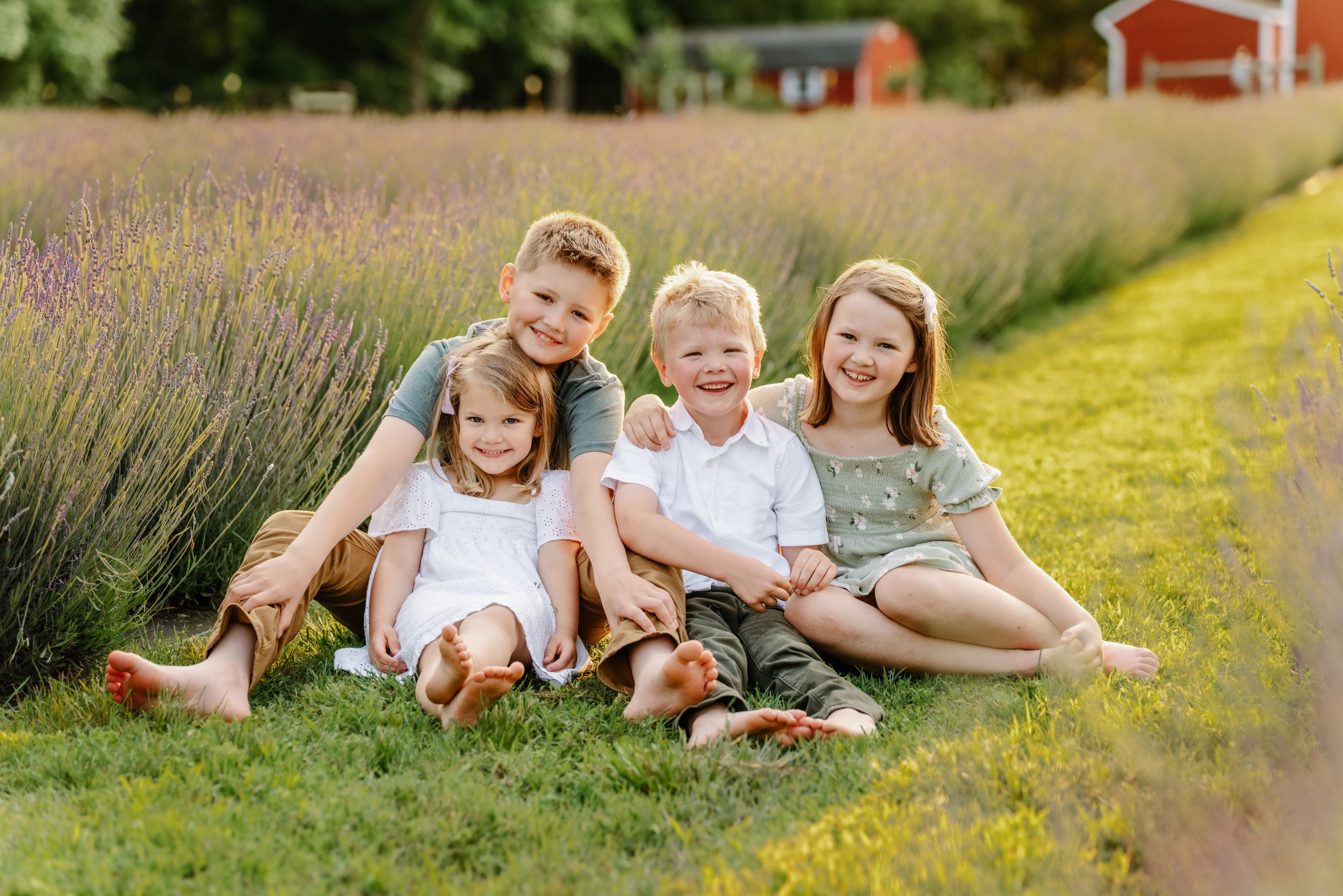 Children sit together in a field of lavender flowers next to a red barn on a sunny summer day.