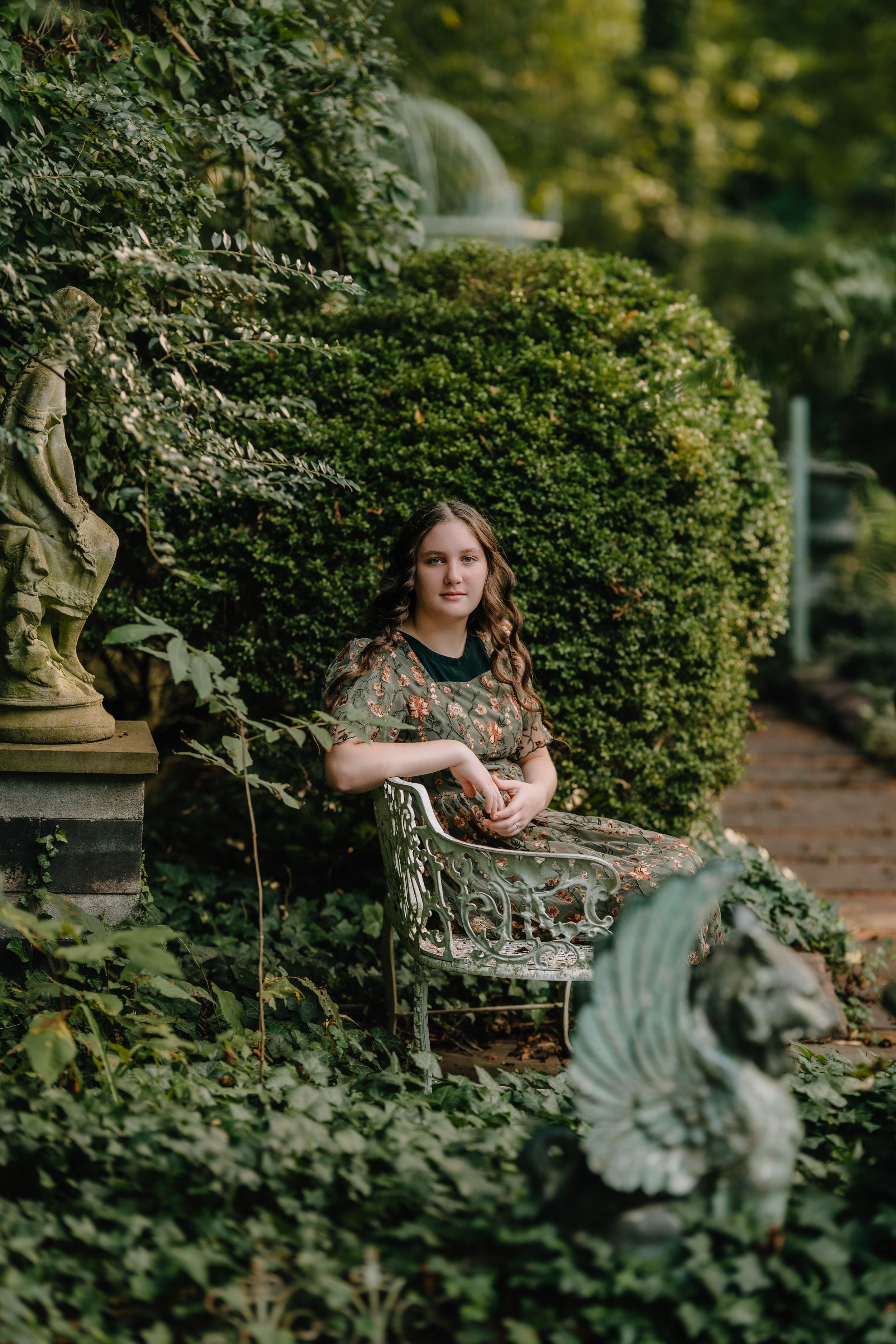 A person sits on an ornate garden bench surrounded by lush green hedges and foliage in a peaceful outdoor setting.