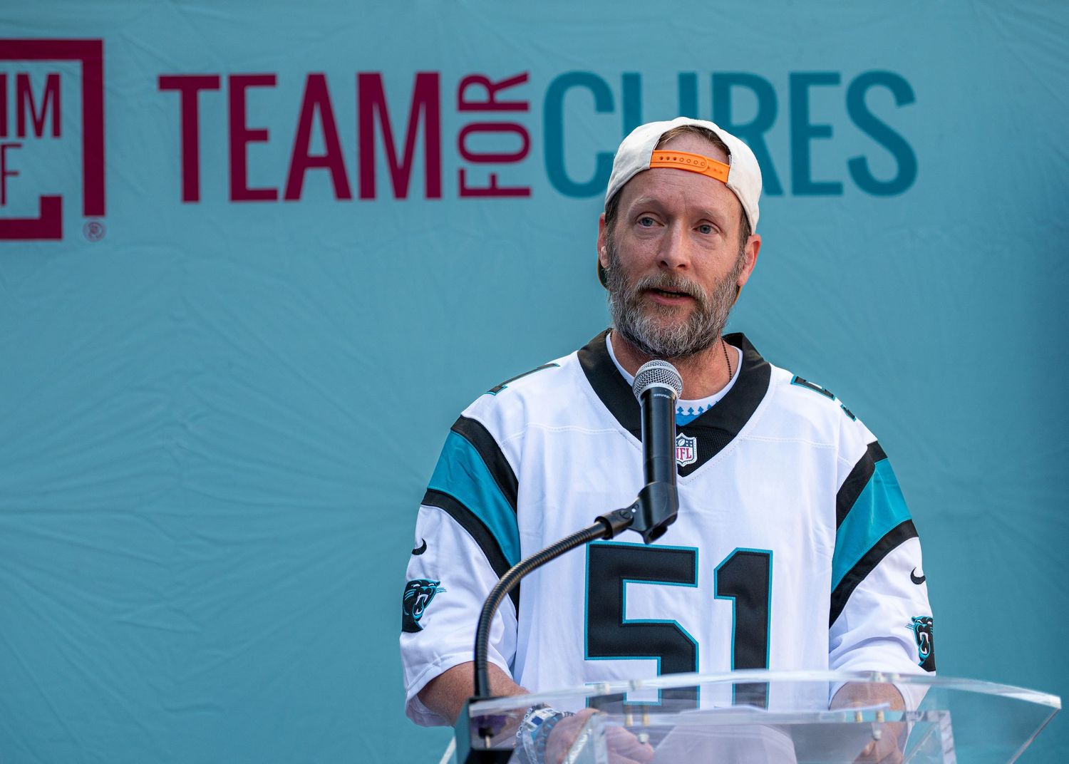 Man in sports jersey #51 speaks at Team for Cures charity event podium outdoors.