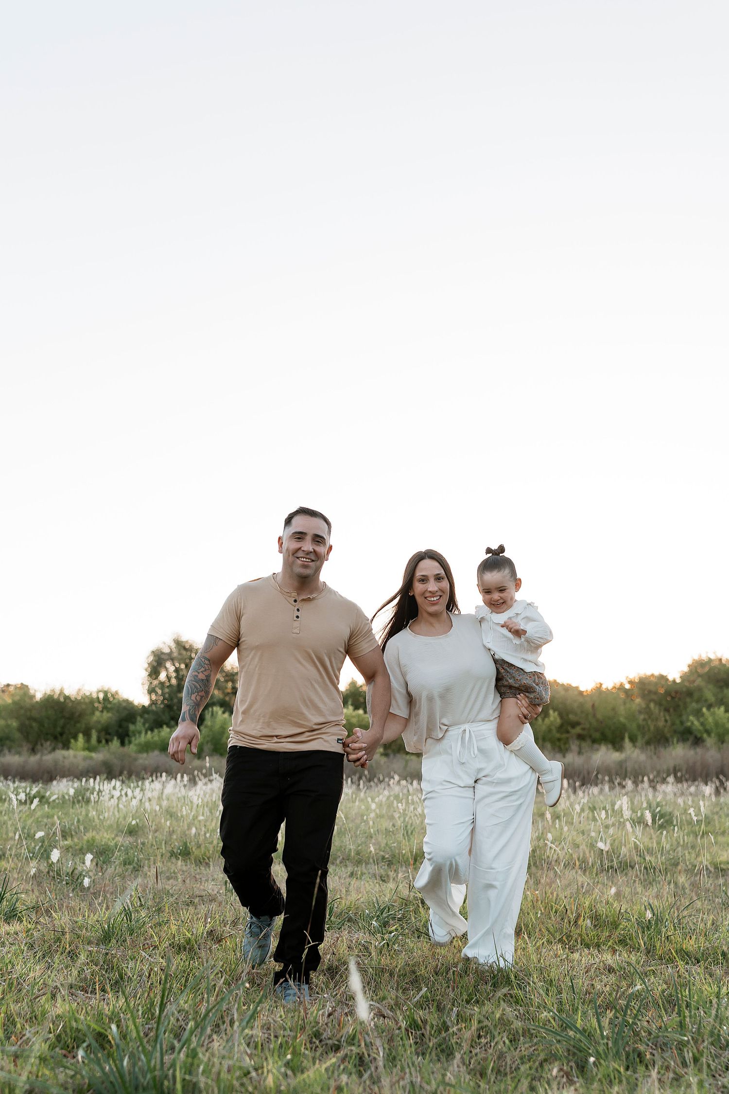 Happy family of three walking in a green field at sunset, parents holding hands with mother carrying toddler.