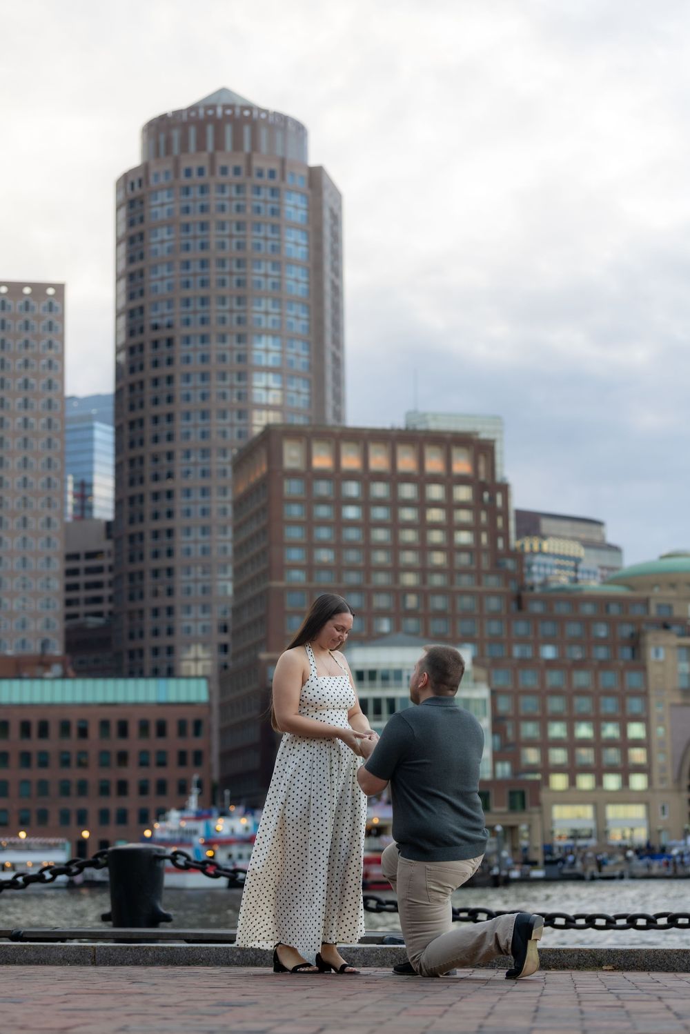 Man proposes on one knee to woman in polka dot dress on Boston waterfront with city skyline backdrop.