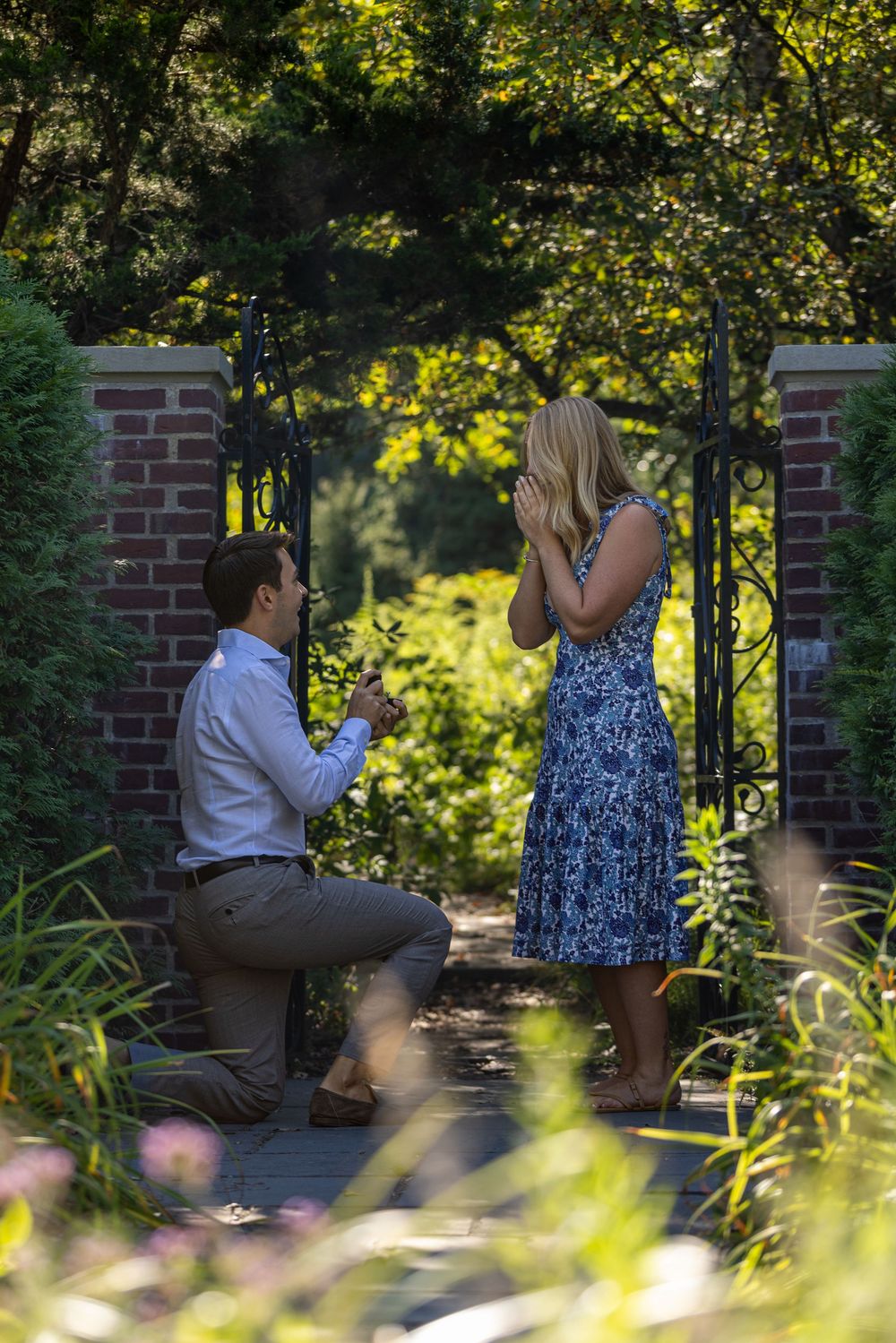 Man proposes on one knee to woman in blue floral dress at a garden gate surrounded by lush greenery.