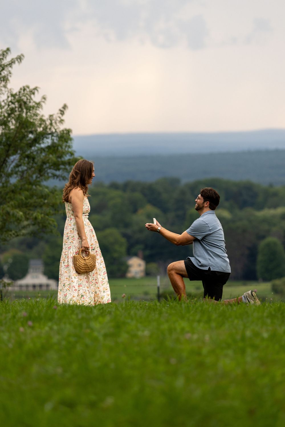 Man proposes on one knee to woman in floral dress on green hillside at sunset.