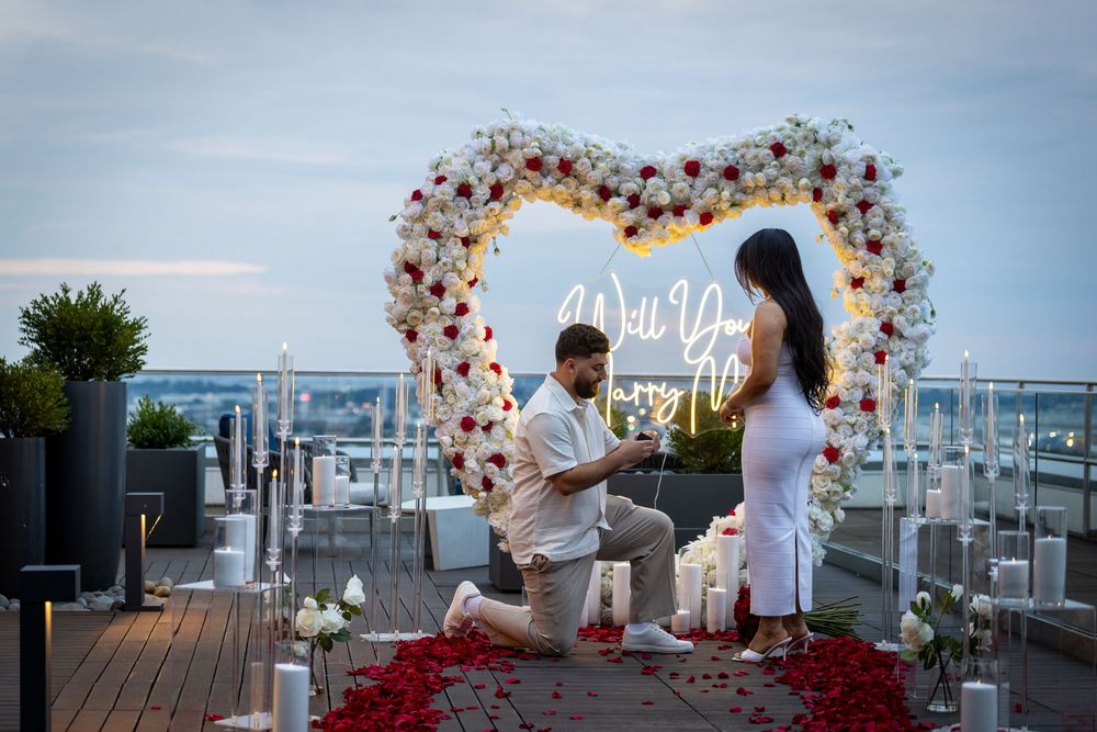 Man proposes on rooftop surrounded by candles, rose petals, and a floral heart arch with a neon Will You Marry Me sign.