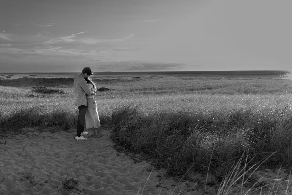 Black and white photo of a couple embracing on a sandy beach path surrounded by tall grass near the ocean.