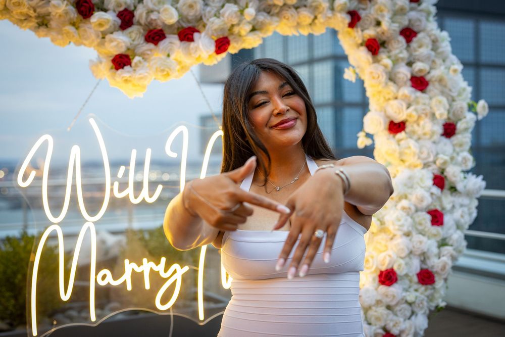 Woman in white dress posing under floral arch with Will You Marry Me neon sign at romantic proposal setup.