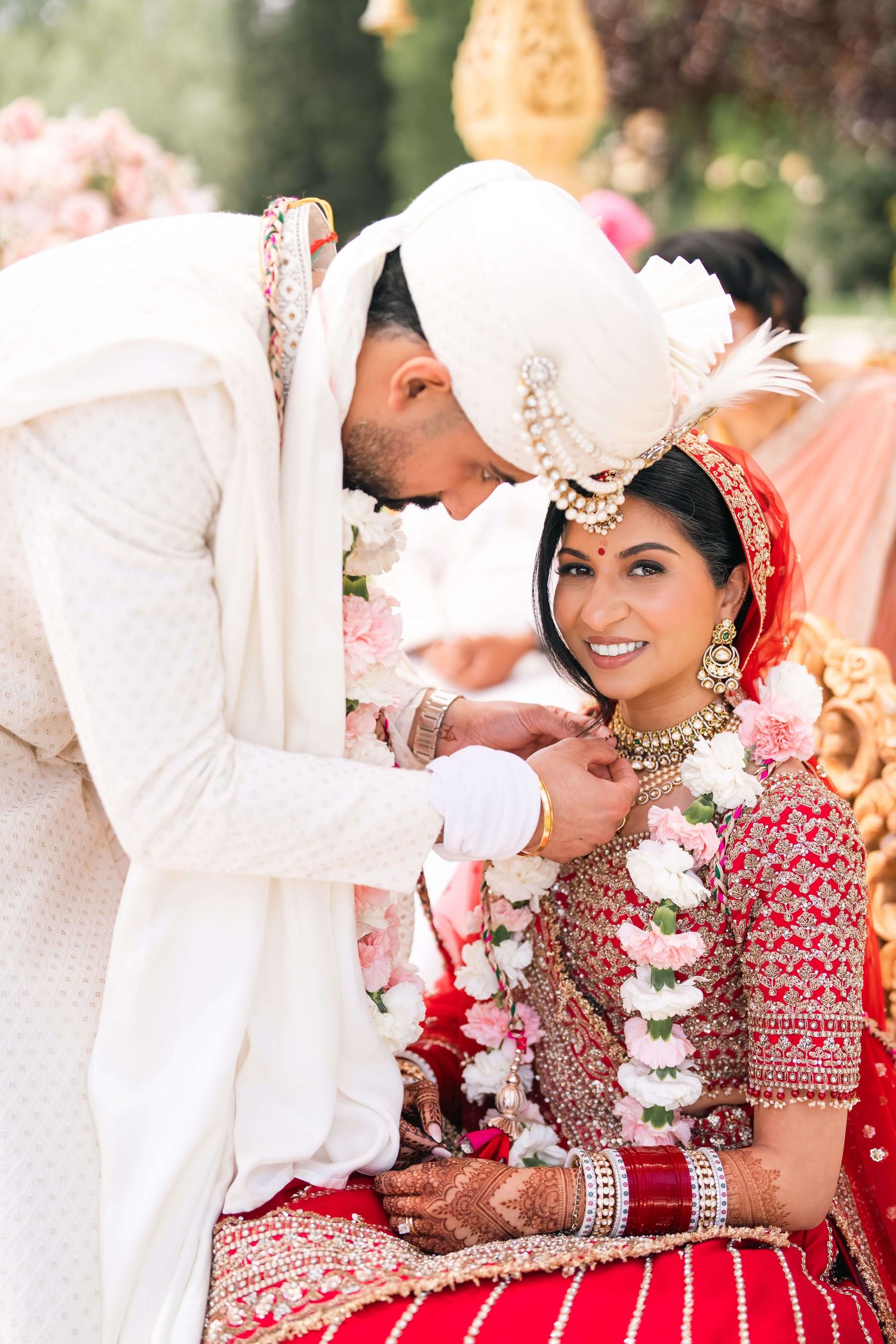 Groom in white sherwani placing garland on smiling bride in red lehenga during traditional Hindu wedding ceremony.