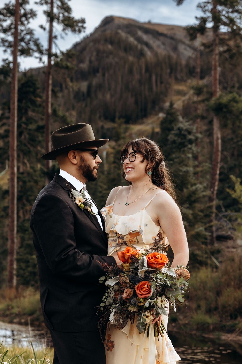 Bride and groom share a romantic moment with mountain backdrop, she holds orange roses bouquet.