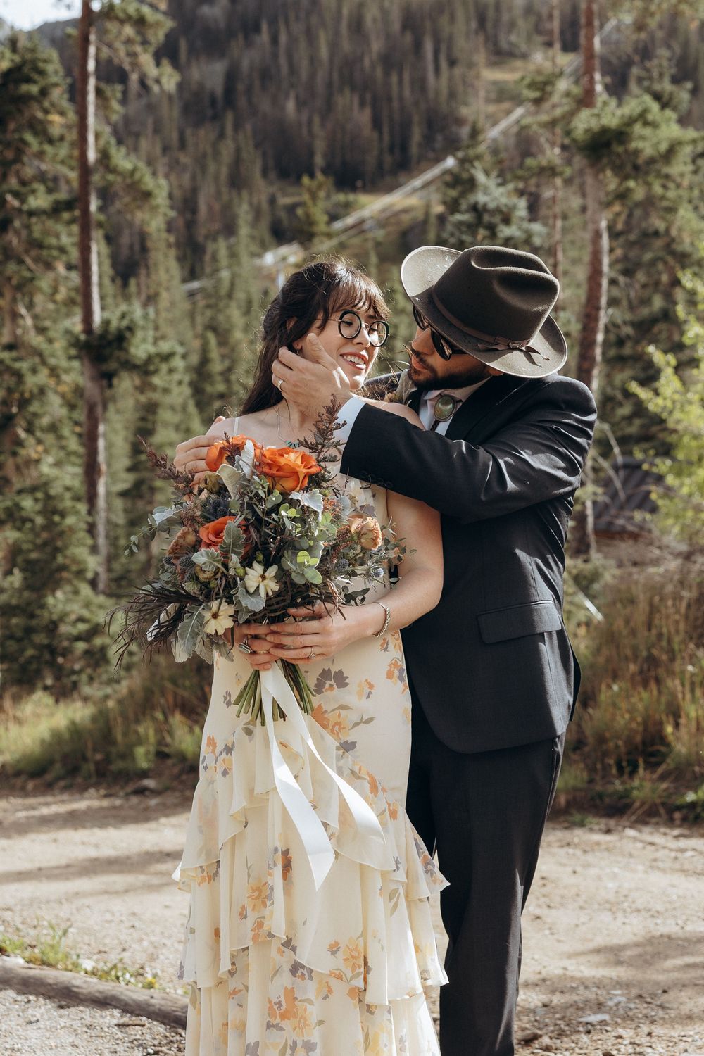 A groom in a black suit and hat tenderly holds his bride's face outdoors, as she holds an autumn bouquet in a floral dress.
