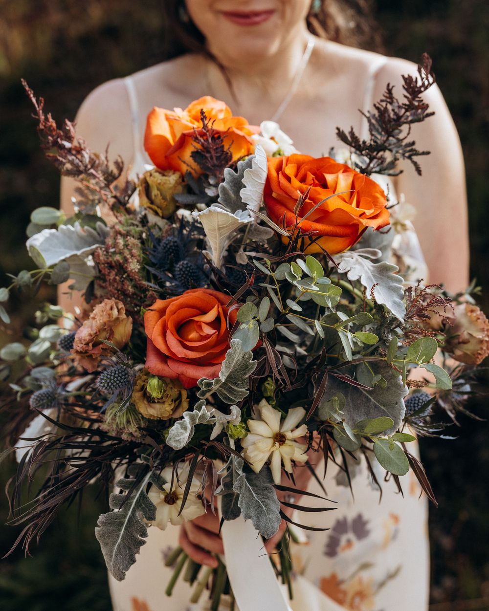 Bride holding a lush autumn wedding bouquet with orange roses, thistle, white daisies, and wild greenery.