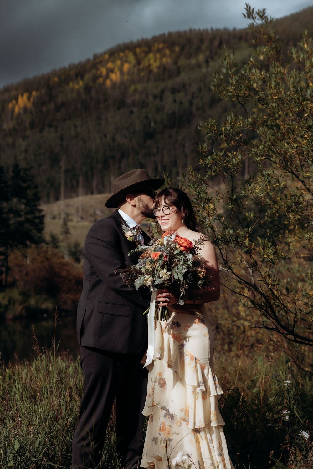 A groom in a black suit and hat kisses his bride holding a bouquet against an autumn mountain backdrop.