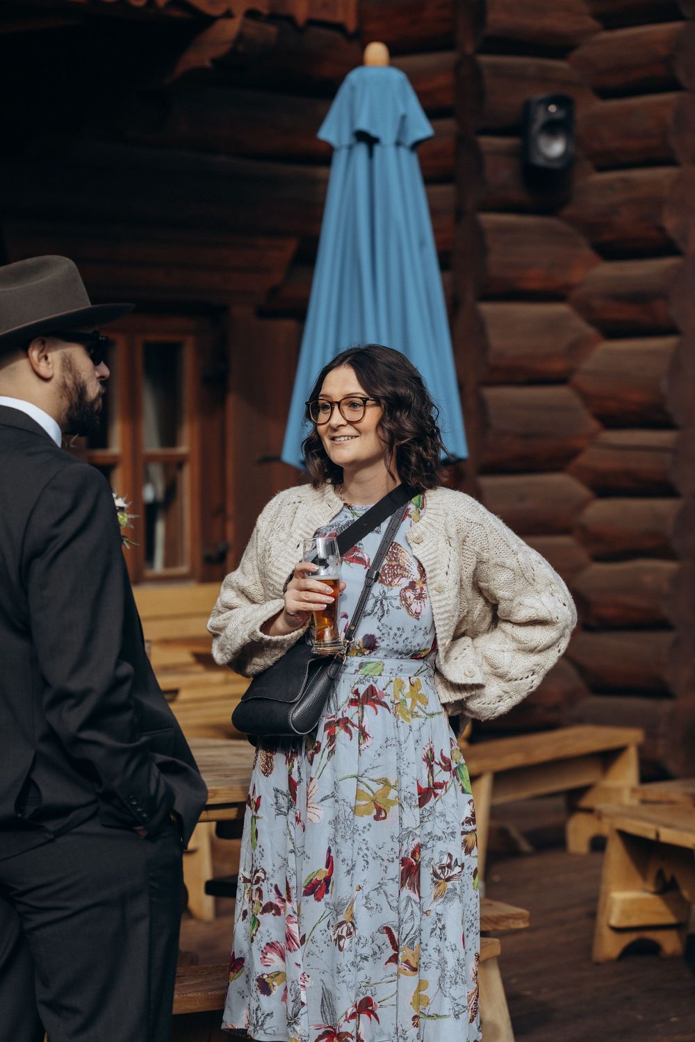 Smiling woman in floral dress holds a drink outside a rustic log cabin wedding venue.