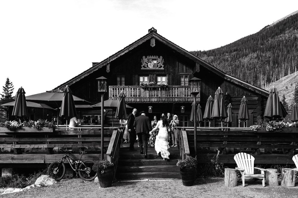 Black and white photo of a wedding couple entering a rustic mountain lodge venue with forested hills in the background.
