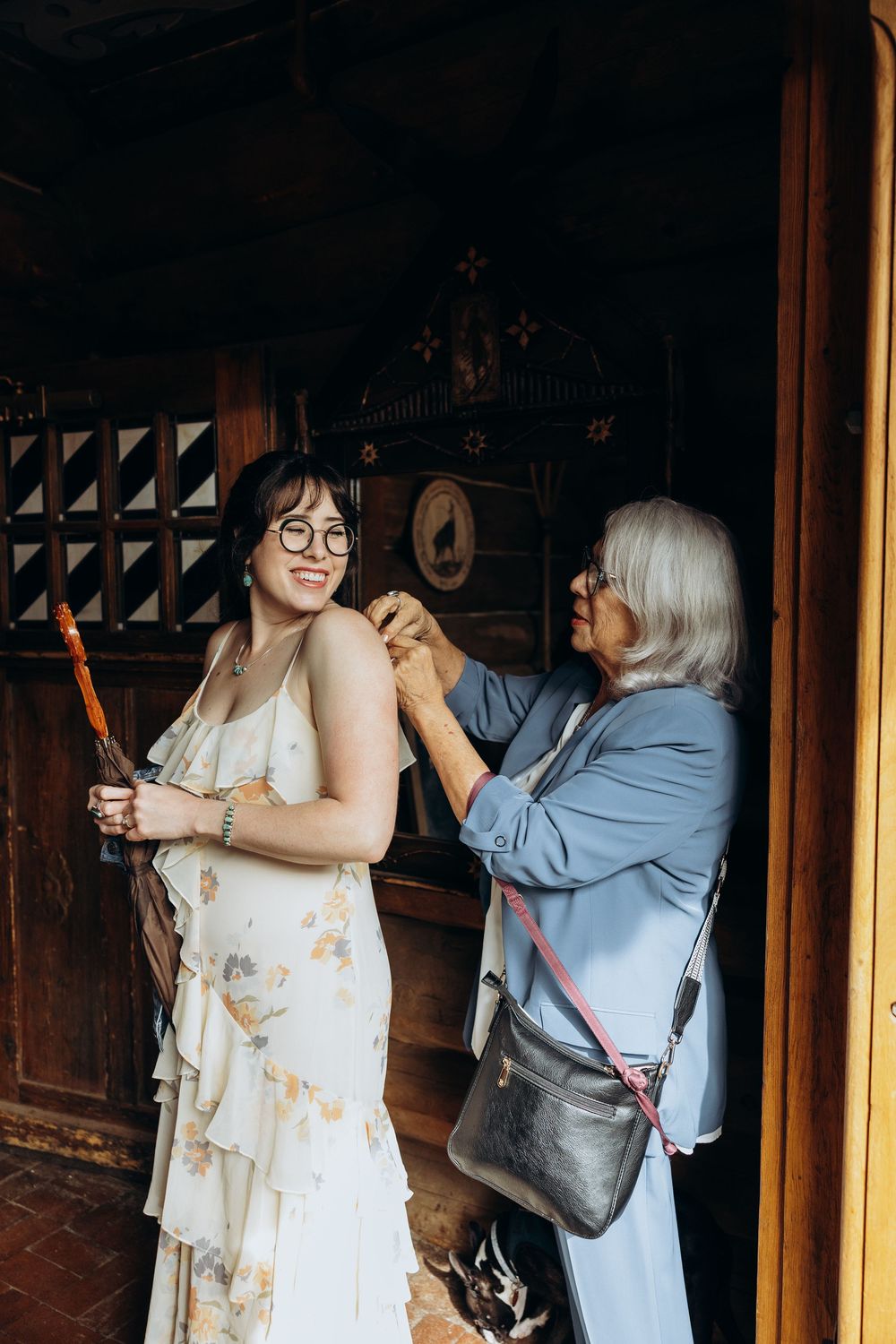 An older woman helps a smiling bride in a floral dress prepare for her wedding inside a rustic wooden venue.