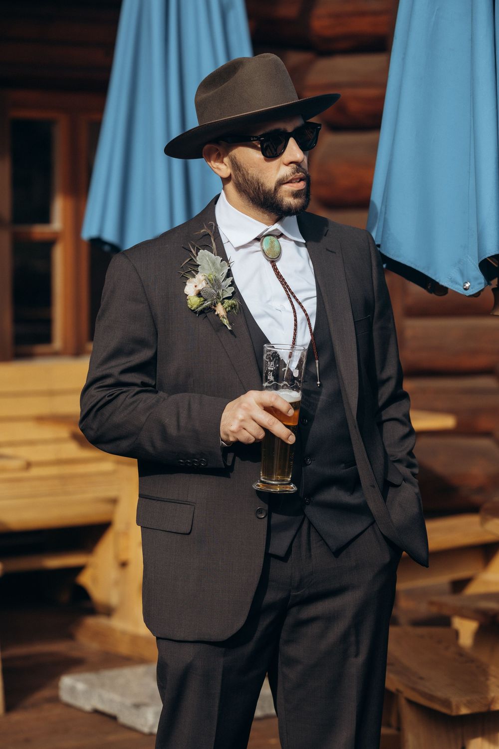 Stylish groom in dark suit, wide-brim hat, and sunglasses holding a beer glass outside a rustic wooden venue.