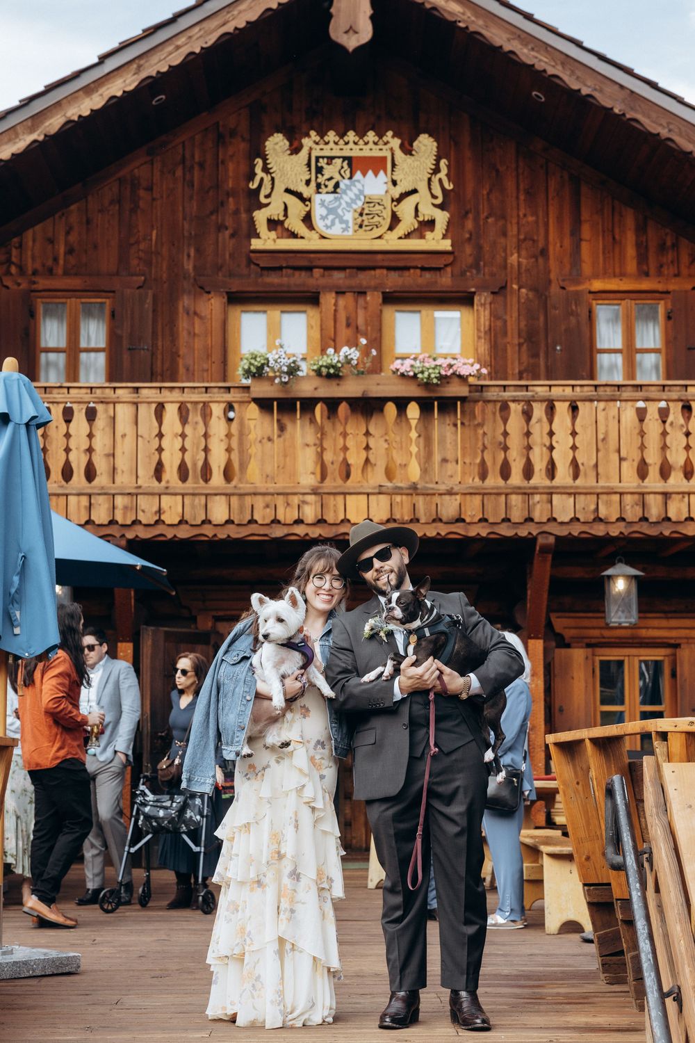 A bride and groom hold their small dogs in front of a traditional wooden Alpine chalet on their wedding day.