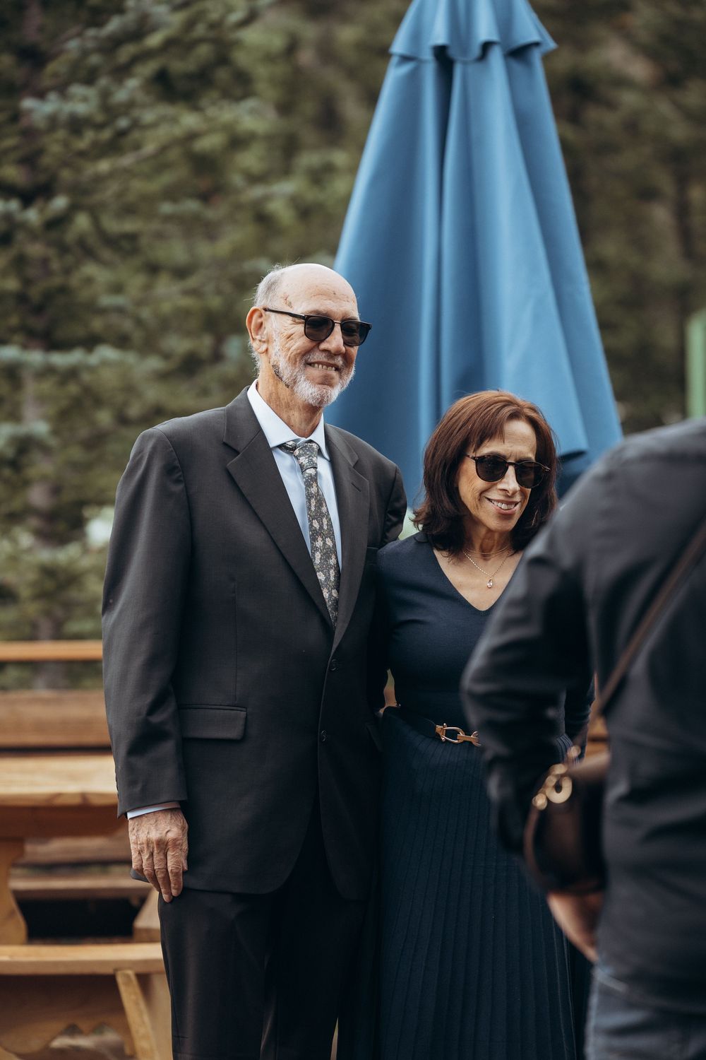 Elegant older couple in sunglasses smile together at an outdoor wedding reception with blue umbrella backdrop.