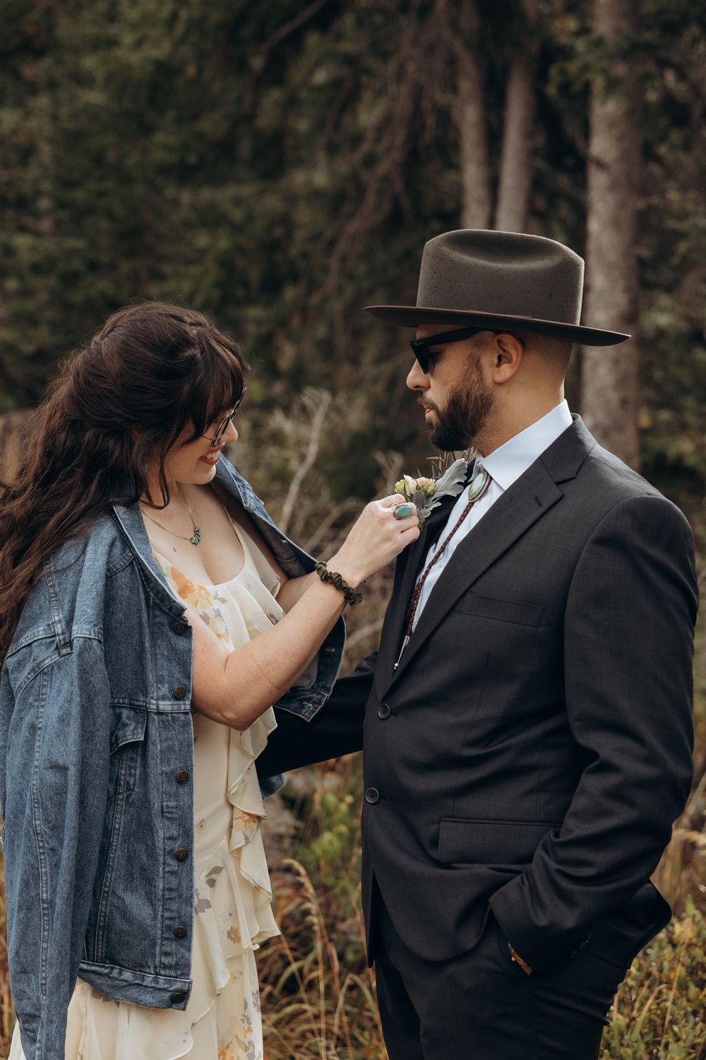Bride in denim jacket pins boutonniere on groom in dark suit and wide-brim hat outdoors.