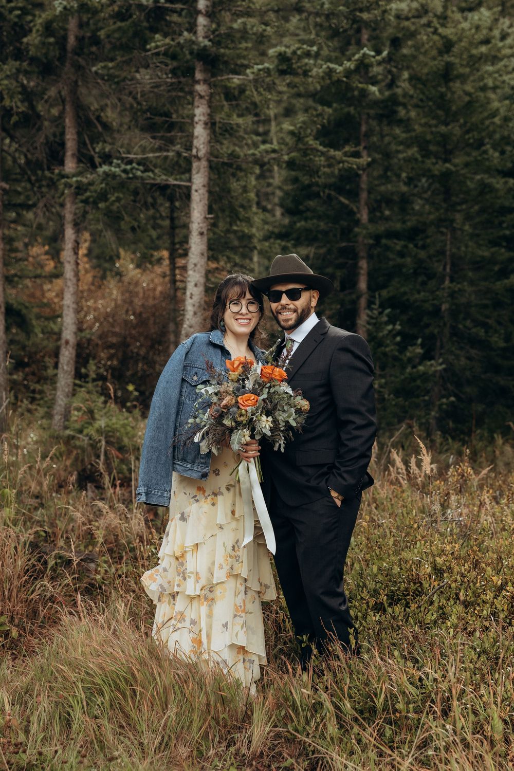 Boho wedding couple posing in forest, bride in floral dress with denim jacket holding orange bouquet, groom in black suit and hat.