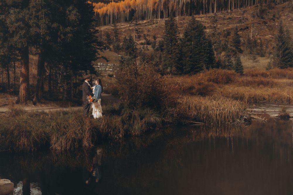 Couple embracing by a reflective mountain lake surrounded by autumn foliage and evergreen trees.