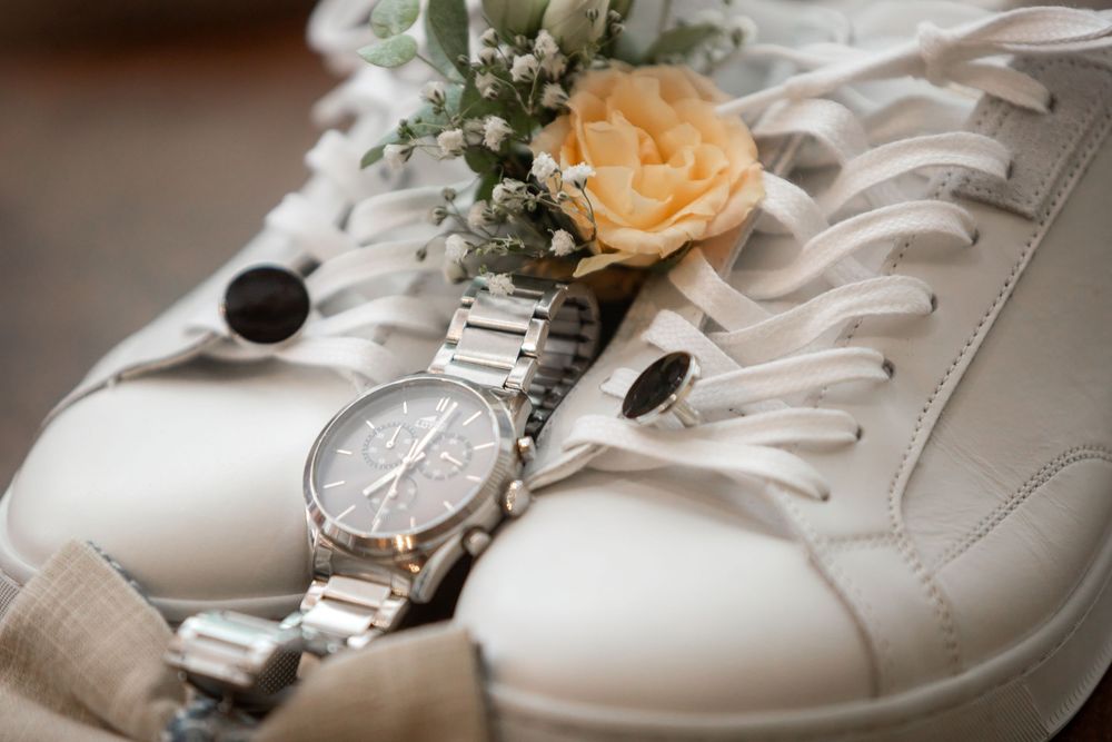 White sneakers with a silver chronograph watch, yellow rose boutonniere, and cufflinks for a stylish groom.