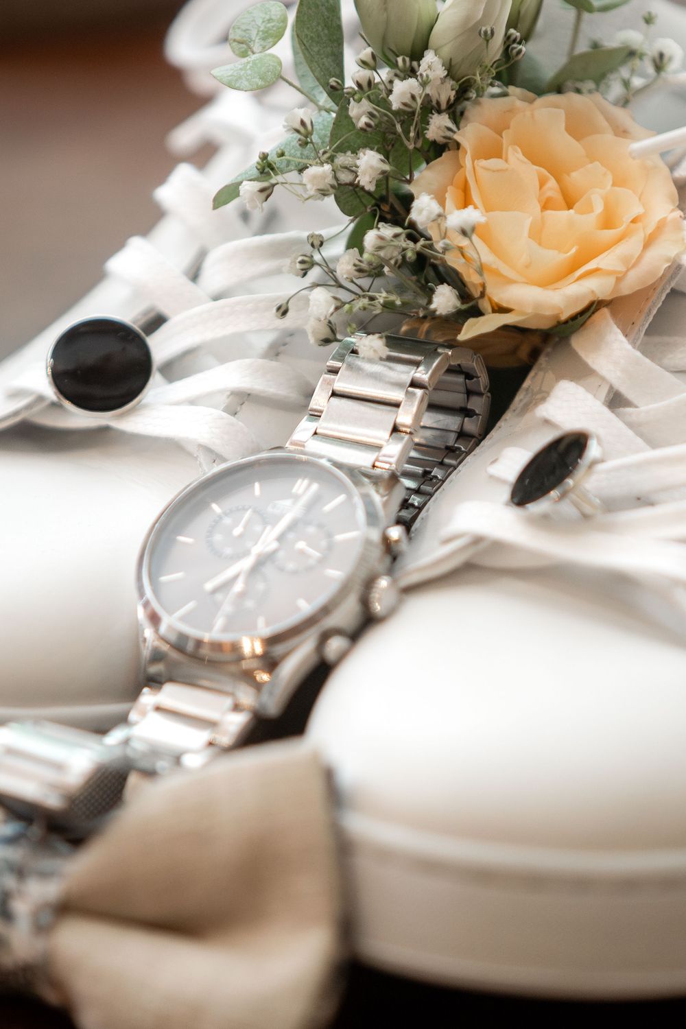 Silver chronograph watch with metal bracelet beside white wedding shoes, cufflinks, and a peach rose boutonniere arrangement.