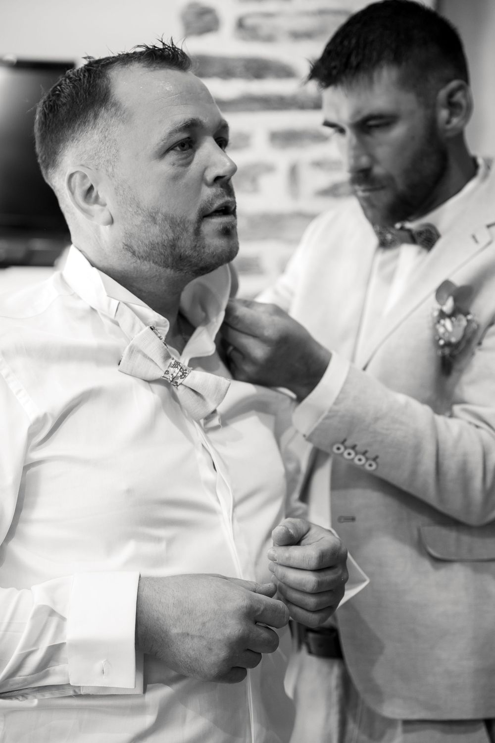 Black and white photo of two men in white suits and bow ties, one helping the other adjust his collar before a wedding.