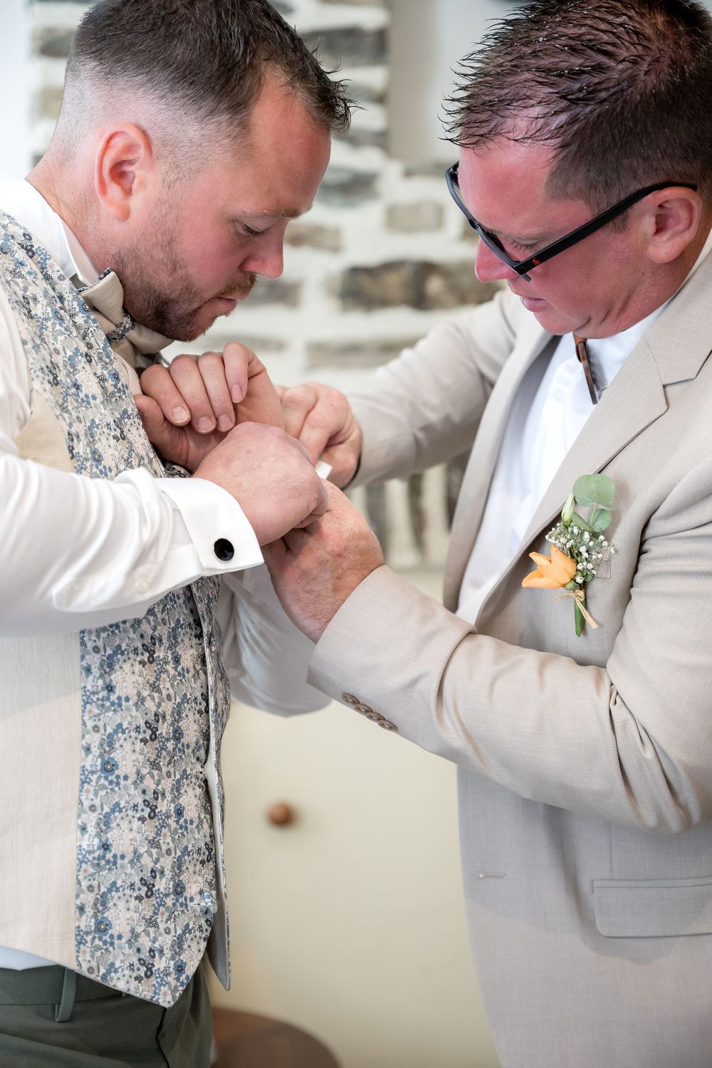 Groom adjusting cufflinks with help from best man in floral vest at wedding ceremony.