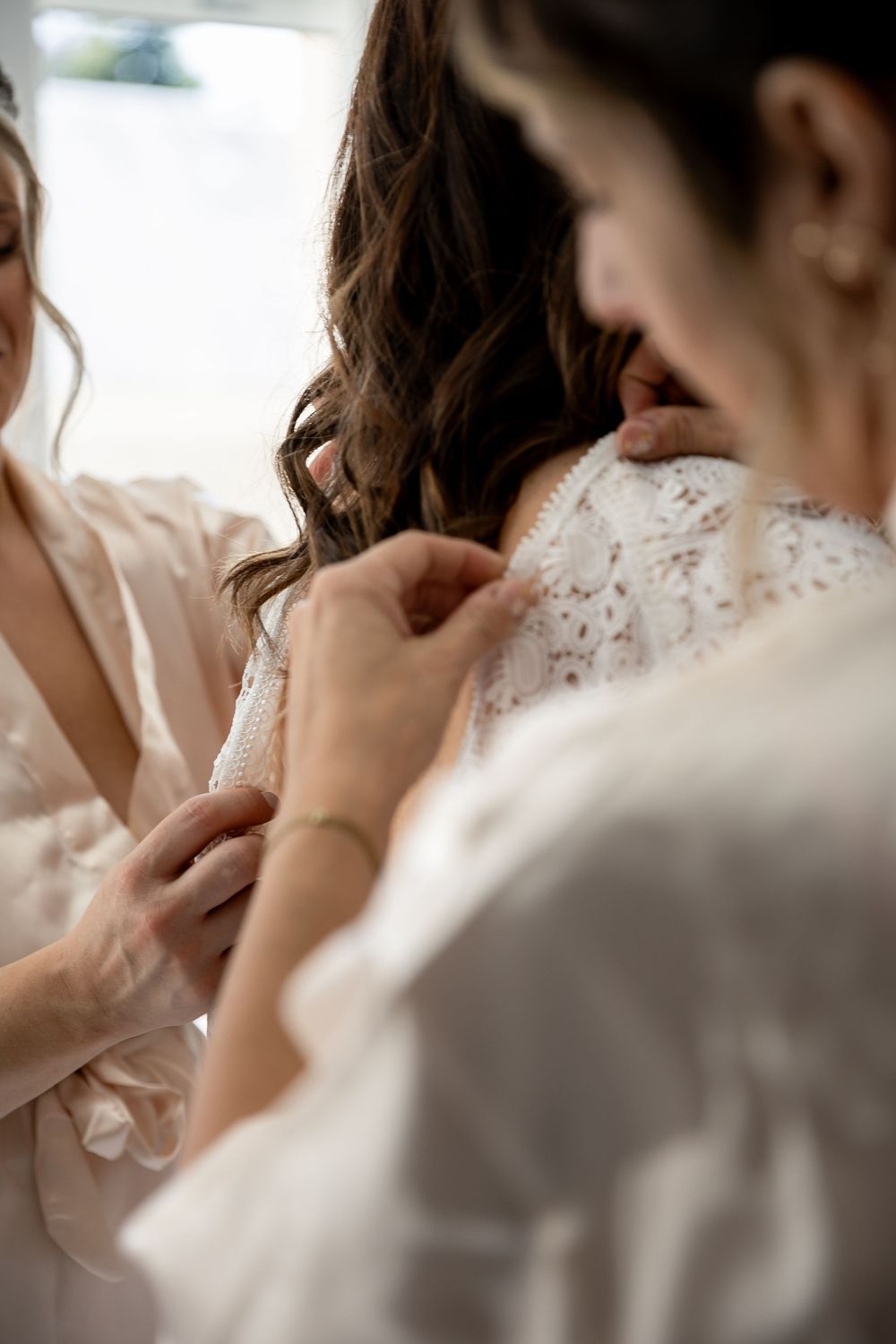 Bridesmaids helping bride button up her lace wedding dress during bridal preparations.