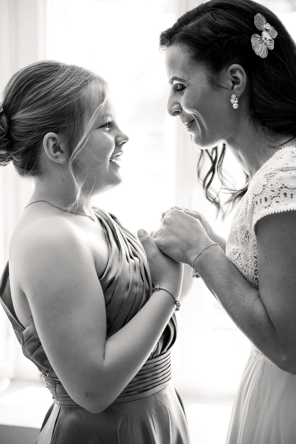 Black and white photo of bride and bridesmaid holding hands and smiling at each other on wedding day.
