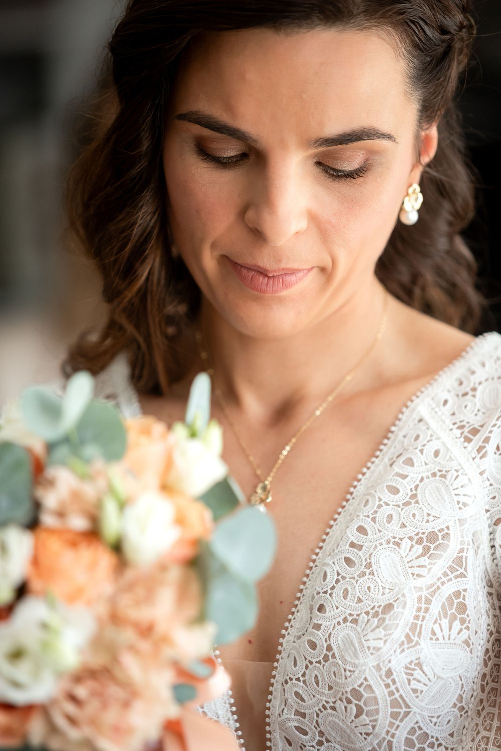 Bride in lace wedding dress holding peach and white bouquet, wearing pearl earrings and gold necklace, looking down softly.