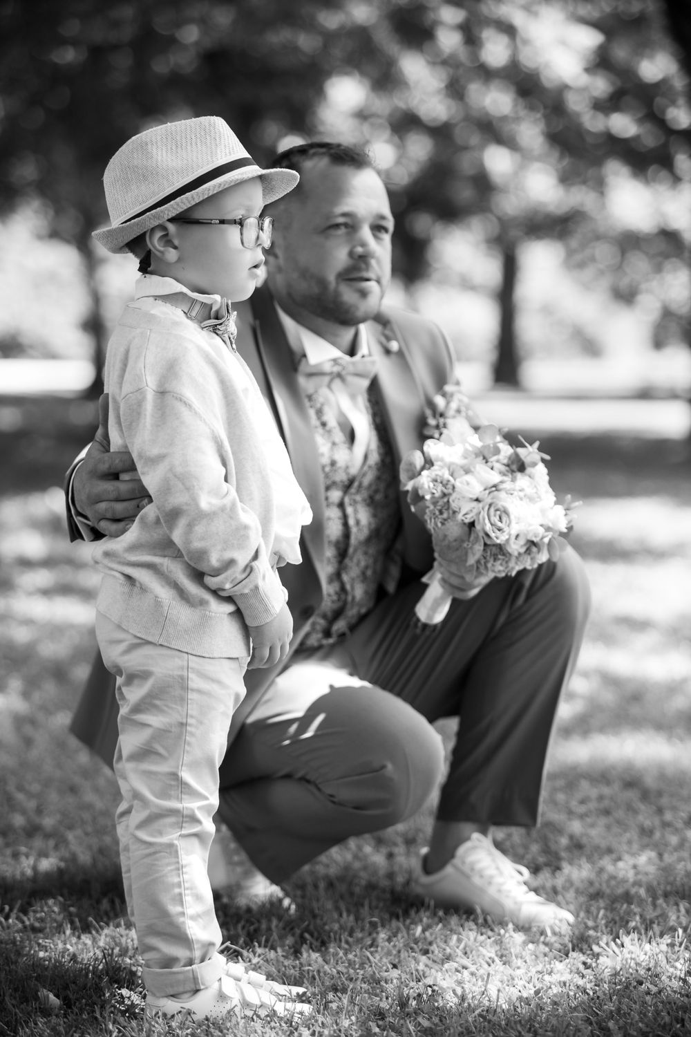Black and white photo of a groom kneeling beside a young boy in a hat, both dressed formally, holding a bouquet outdoors.