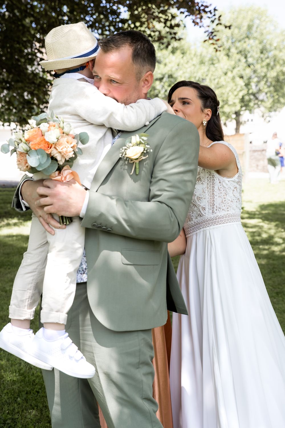 Groom in sage suit hugs child while bride in white gown looks on during outdoor wedding ceremony.
