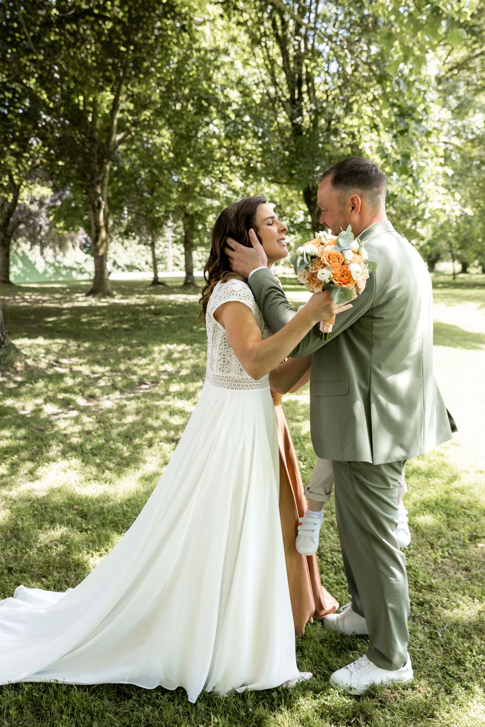 A bride in a white gown and groom in a sage suit share a romantic moment in a sunlit park holding orange flowers.