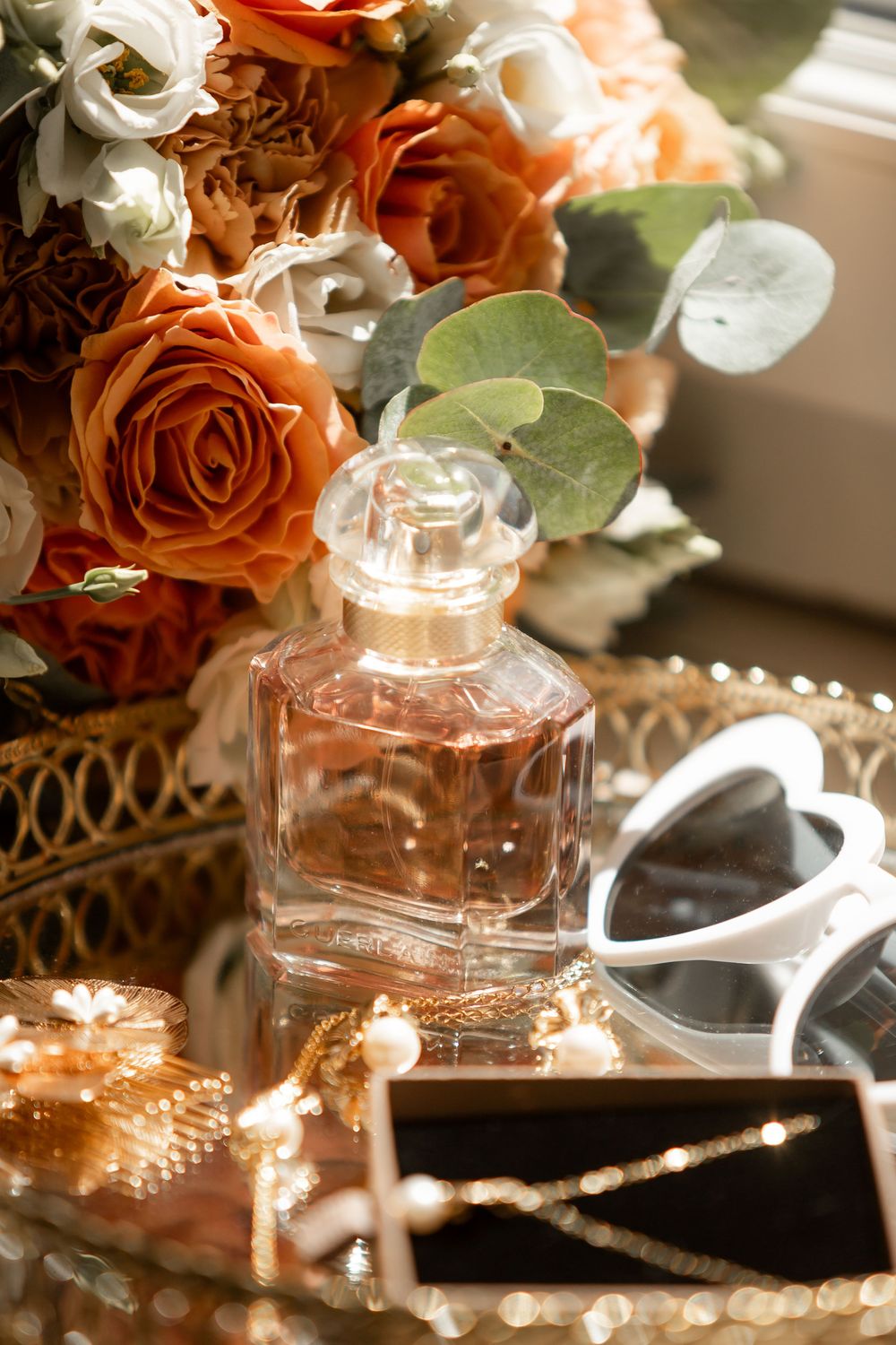 Elegant perfume bottle on a mirrored tray with sunglasses, jewelry, and orange roses bouquet in warm sunlight.