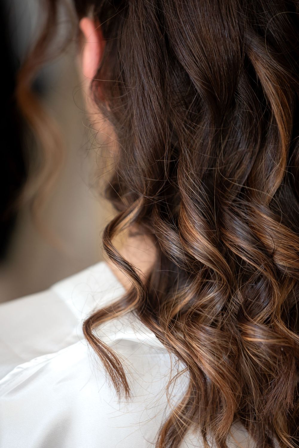 Close-up of woman's dark brown curly hair with caramel highlights, worn down over a white shirt.