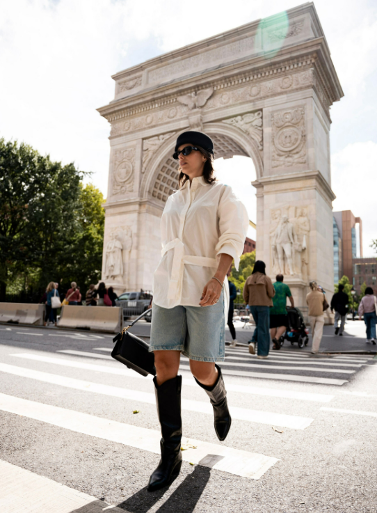 Person in white belted shirt, denim shorts, black boots and cap at Washington Square Park arch.