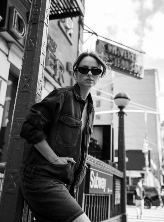 Woman in denim jacket and sunglasses posing on urban street near Subway sign, black and white photo.