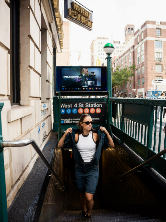 Woman emerging from West 4th St Station subway entrance in NYC, wearing white top, denim skirt, and sunglasses.