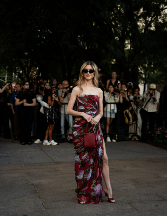 Woman in floral strapless maxi dress with slit poses outdoors at fashion event with photographers in background.